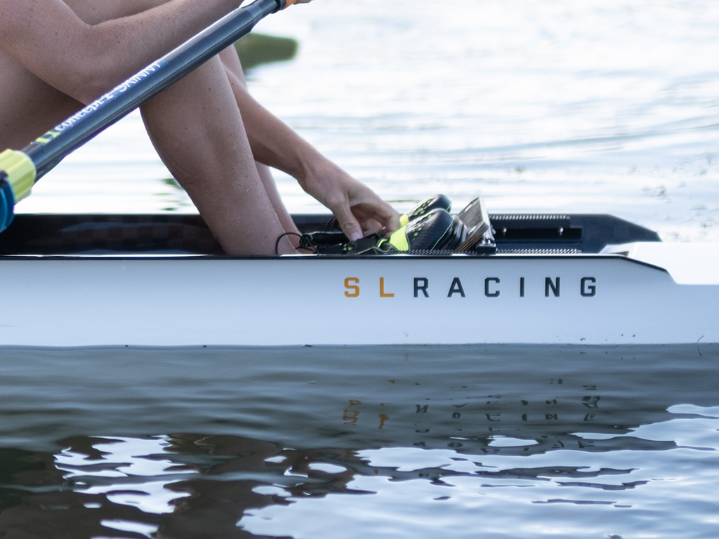 An SL Racing single scull sits on the water, a rower adjusts her feet in the shoes in the boat.