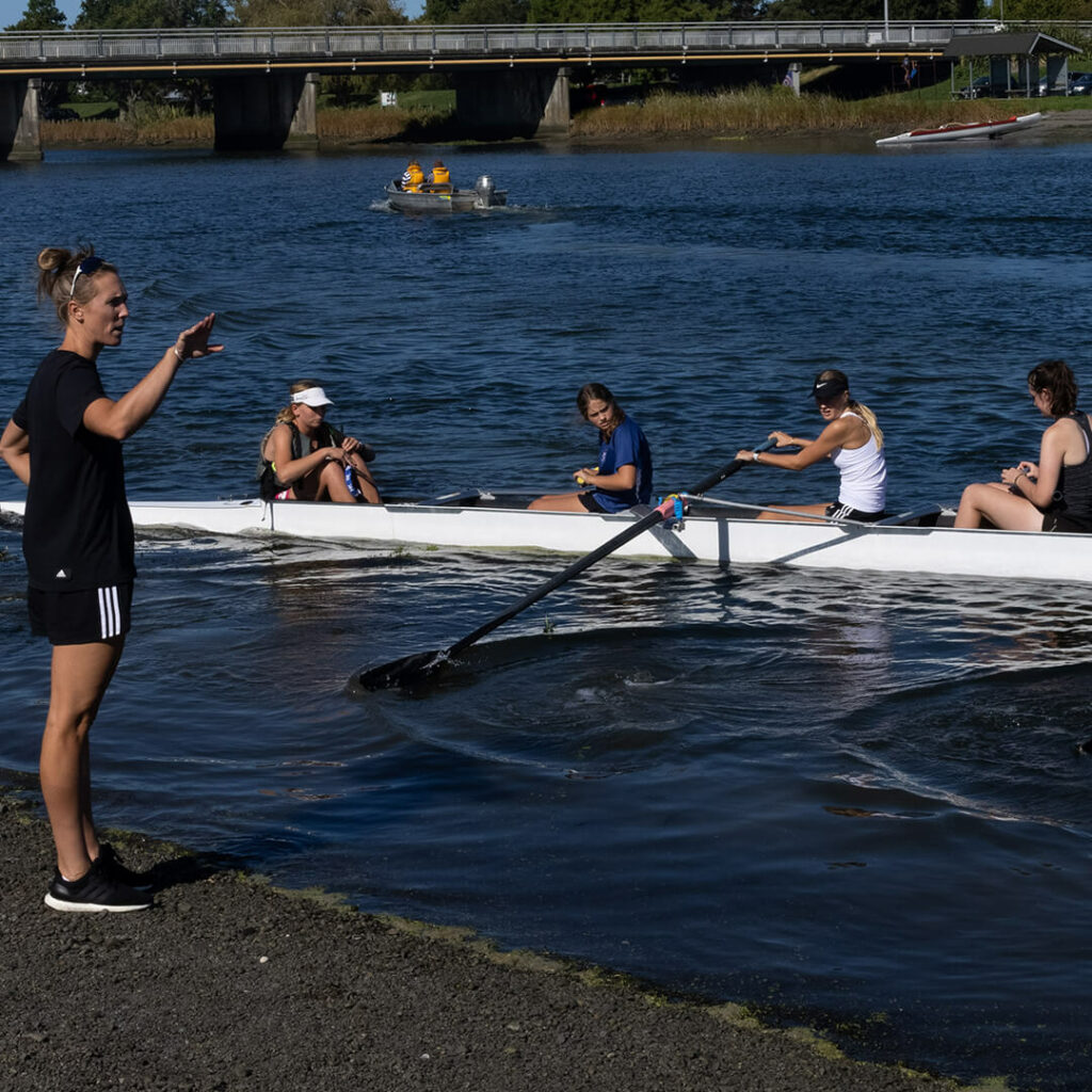 Emma Twigg stands on the banks of the Clive River coaching a group of teenage girls in an eight rowing skiff