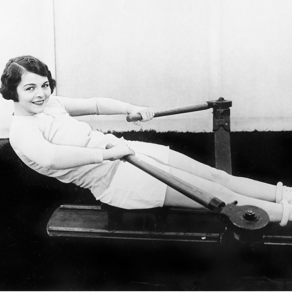 An old black and white photo of a women in white shorts and a white long sleeve top on an old rowing machine