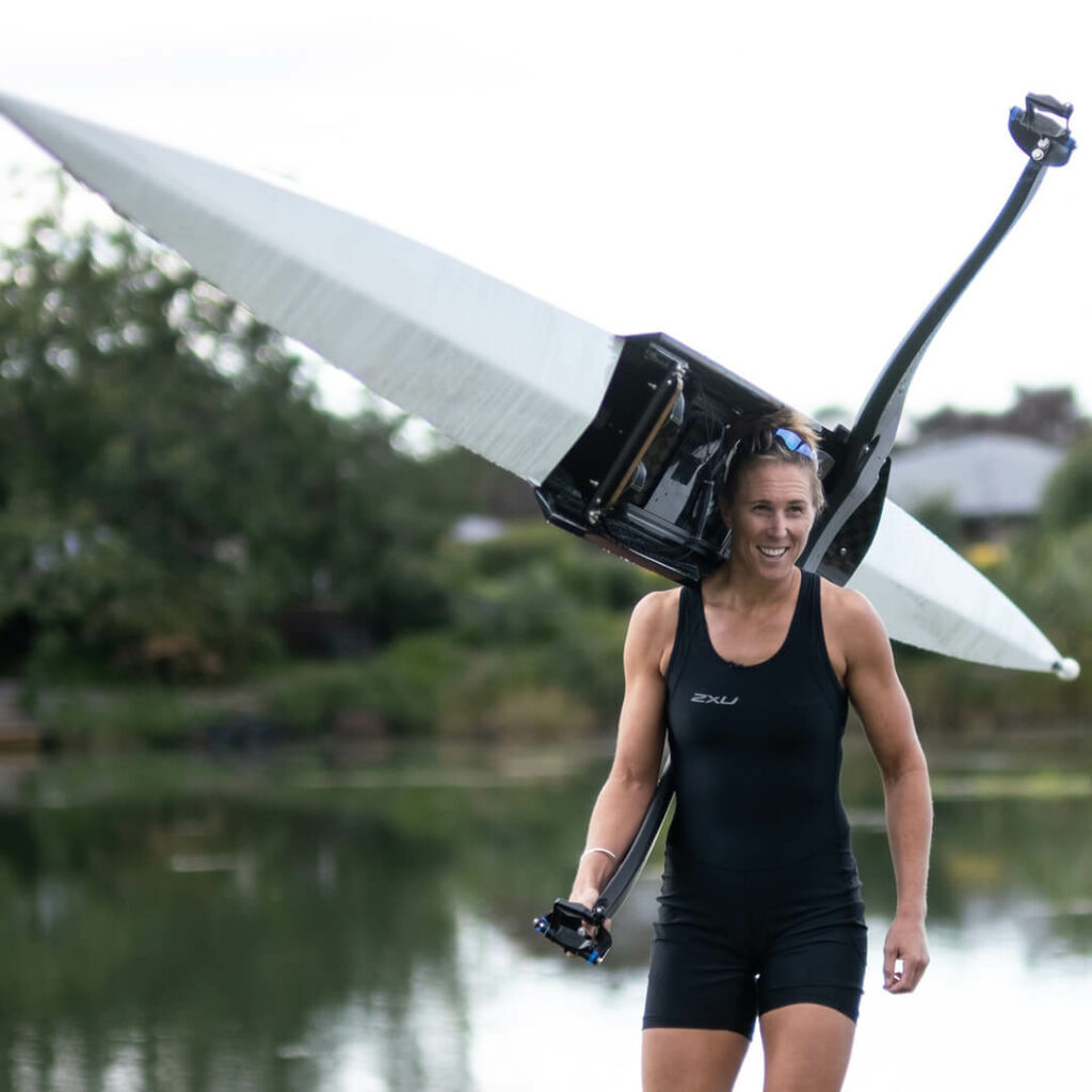 Olympic champion rower carries a single SL Racing skiff over her shoulder as she walks up the bank of the River Clive