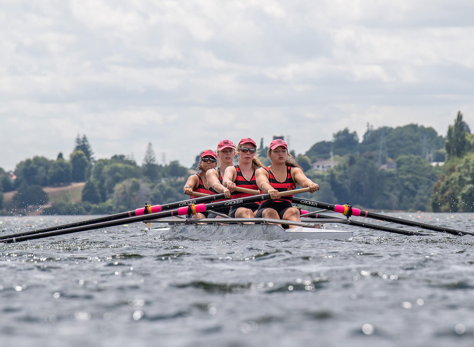 The camera is low in the water. There are four girls in red and black stripped tops with pink hats racing in the water on a S L racing coxless four quad rowing boat. They have determined looks on their faces.
