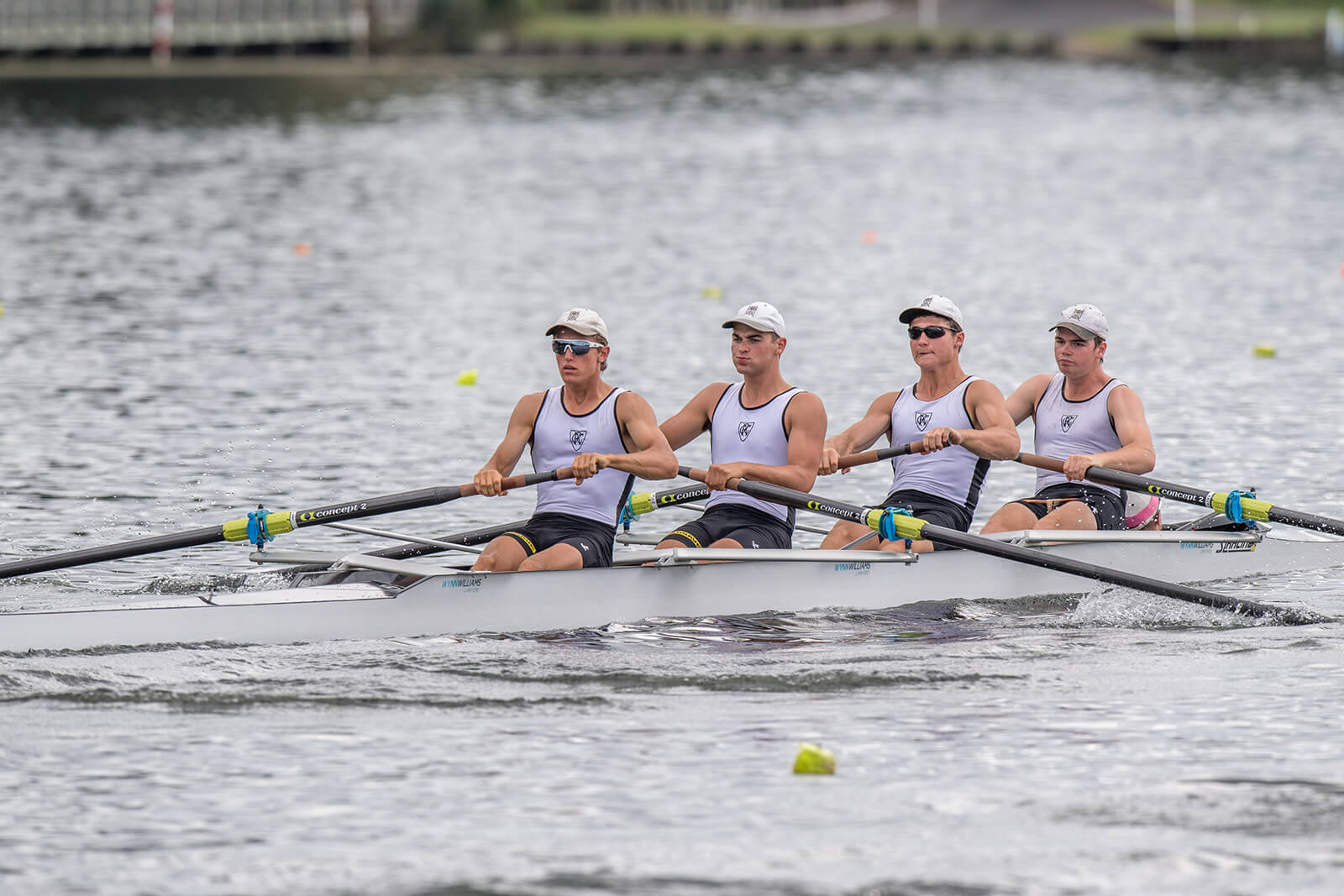 There are four young men racing an SL Racing coxless four rowing boat on the river. They have black oars and are rowing in sync, wearing white tops and white caps.