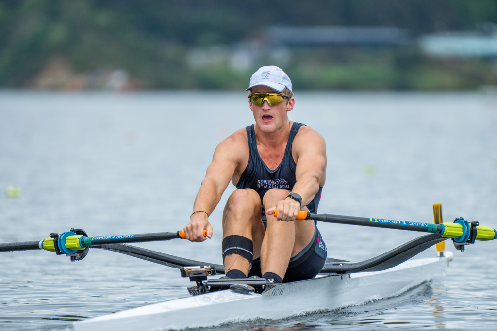 Ben is rowing on an SL racing single scull racing boat, facing the camera, holding onto his oars on a lake with a blurred background behind hin.