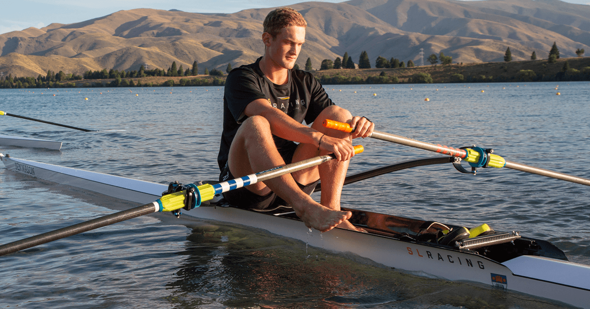 Ben sits on a single scull SL Racing rowing boat. The lake is clear and there is a mountain range behind him. He is wearing a black t-shirt and holding oars.