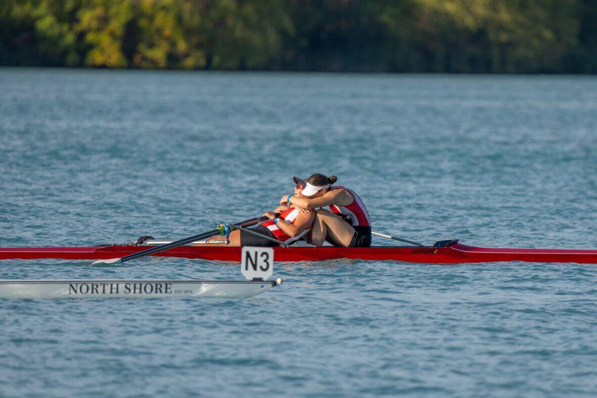 Two female rowers celebrate their finish on the water. The girl in front is leaning back as the girl behind her reaches her arms around her neck to hug her. They are in red and white stripped tops in a red double skiff.