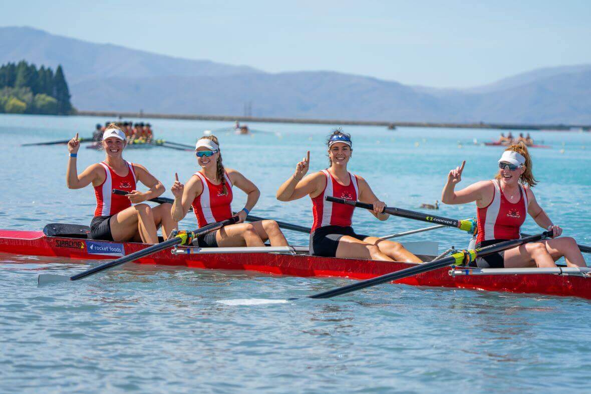 Four young female rowers hold up their index fingers to represent 'one' at a NZ rowing competition in a red coxless four quad SL Racing boat