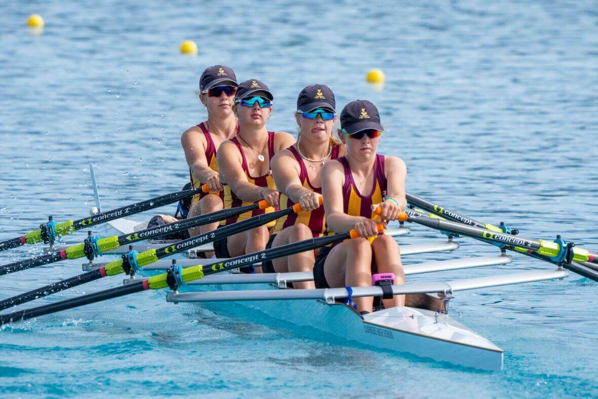 Four young female rowers compete in an SL Racing coxless four quad rowing boat. They wear maroon and yellow stripped tops, blue caps and reflective sunglasses. They have looks of intense concentration on their faces.