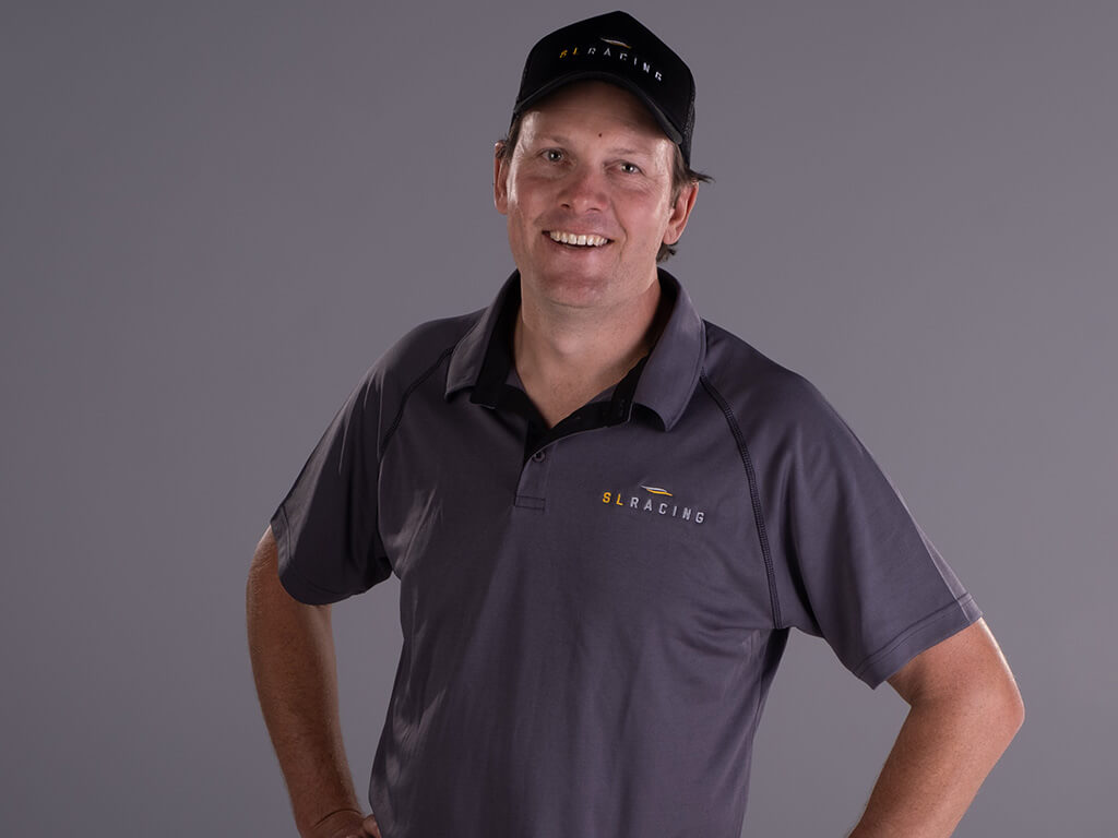 S L Racing owner and founder Simon Lack stands with his hands on his hips wearing a black S L Racing cap and a grey SL Racing shirt on a grey backgroud.